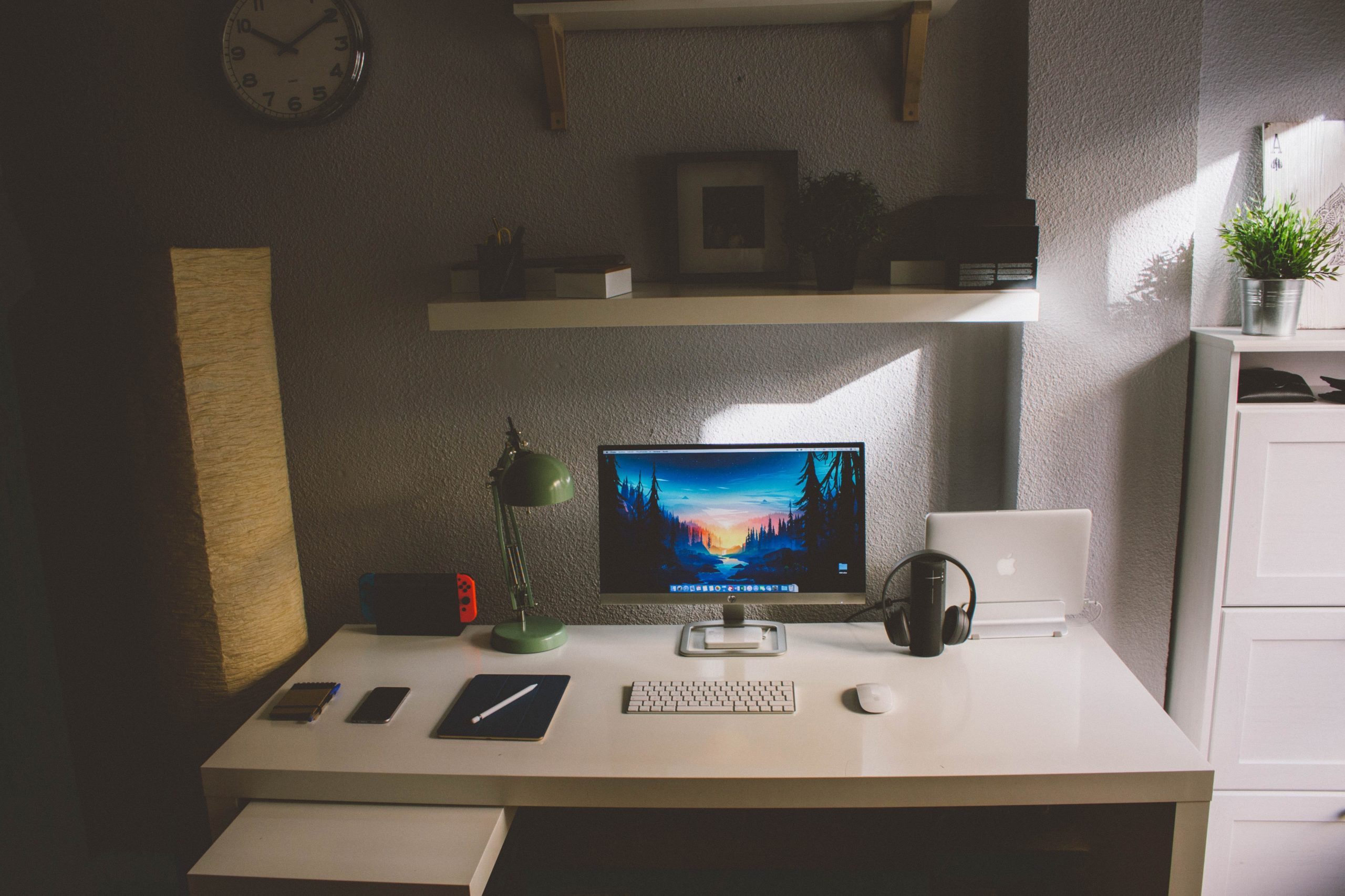 A dimly lit photo of a sparse working desktop with a monitor, keyboard, notebooks and a beam of light from a nearby window