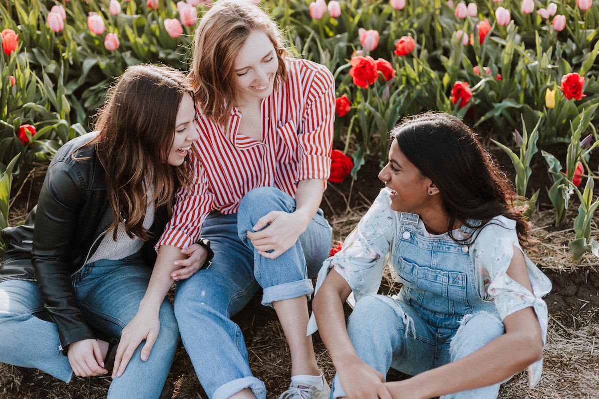Three women lauging together harmoniously in a sunlit meadow of flowers.